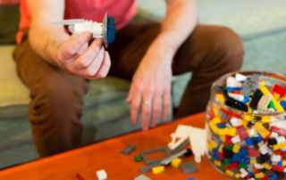 A person holding up a creation made from Lego bricks, with a table strews with Legos in front of them.