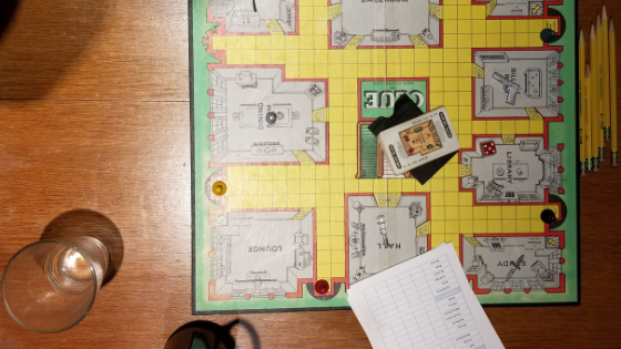 photo of a vintage Clue board game game board on a woodgrain table with game cards, pencils and beverage glasses around.